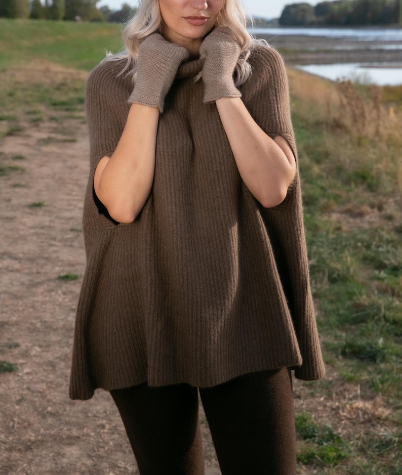 May include: A brown ribbed knit poncho with a high turtleneck collar and matching brown knit gloves. The garment features an open-side design with arm openings, providing a loose, flowing fit that extends to the upper thighs in a solid earth tone.