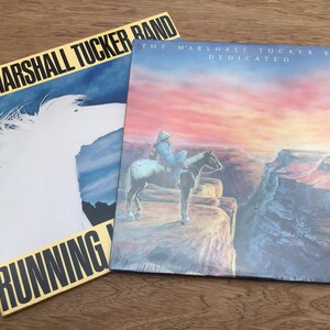 May include: Two vinyl records by The Marshall Tucker Band. The first record is titled "Running..." and features a white horse on a blue and white background. The second record is titled "Dedicated" and features a sunset over a canyon with a person riding a horse.