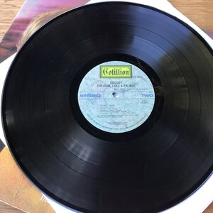 May include: Black vinyl record with a white label. The label has the text "Cotillion" in a yellow box, "TRILOGY", "EMERSON, LAKE & PALMER", "STEREO", "TWO", and "SIDE TWO" printed on it.
