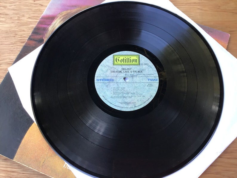 May include: Black vinyl record with a white label. The label has the text "Cotillion" in a yellow box, "TRILOGY", "EMERSON, LAKE & PALMER", "STEREO", "TWO", and "SIDE TWO" printed on it.