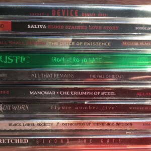 Może przedstawiać: Stos dziewięciu płyt kompaktowych (CD) z r&oacute;żnymi nazwami zespoł&oacute;w i tytułami album&oacute;w. Płyty CD znajdują się w plastikowych pudełkach w kolorze srebrnym i czarnym. G&oacute;rna płyta CD jest czarna z nazwą zespołu "DEVICE" i tytułem albumu "SALIVA BLOOD STAINED LOVE STORY". Druga płyta CD jest czarna z nazwą zespołu "ALL SHALL PERISH" i tytułem albumu "THE PRICE OF EXISTENCE". Trzecia płyta CD jest zielona z nazwą zespołu "CALISTIC" i tytułem albumu "FROM ZERO TO HATE". Czwarta płyta CD jest czarna z nazwą zespołu "ALL THAT REMAINS" i tytułem albumu "THE FALL OF IDEALS". Piąta płyta CD jest czarna z nazwą zespołu "MANOWAR" i tytułem albumu "THE TRIUMPH OF STEEL". Sz&oacute;sta płyta CD jest czarna z nazwą zespołu "OIL WORK" i tytułem albumu "FIGURE NUMBER FIVE". Si&oacute;dma płyta CD jest czarna z nazwą zespołu "BLACK LABEL SOCIETY" i tytułem albumu "CATACOMBS OF THE BLACK VATICAN". &Oacute;sma płyta CD jest czerwona z nazwą zespołu "WRETCHED" i tytułem albumu "BEYOND THE GATE".