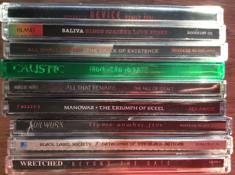 Może przedstawiać: Stos dziewięciu płyt kompaktowych (CD) z r&oacute;żnymi nazwami zespoł&oacute;w i tytułami album&oacute;w. Płyty CD znajdują się w plastikowych pudełkach w kolorze srebrnym i czarnym. G&oacute;rna płyta CD jest czarna z nazwą zespołu "DEVICE" i tytułem albumu "SALIVA BLOOD STAINED LOVE STORY". Druga płyta CD jest czarna z nazwą zespołu "ALL SHALL PERISH" i tytułem albumu "THE PRICE OF EXISTENCE". Trzecia płyta CD jest zielona z nazwą zespołu "CALISTIC" i tytułem albumu "FROM ZERO TO HATE". Czwarta płyta CD jest czarna z nazwą zespołu "ALL THAT REMAINS" i tytułem albumu "THE FALL OF IDEALS". Piąta płyta CD jest czarna z nazwą zespołu "MANOWAR" i tytułem albumu "THE TRIUMPH OF STEEL". Sz&oacute;sta płyta CD jest czarna z nazwą zespołu "OIL WORK" i tytułem albumu "FIGURE NUMBER FIVE". Si&oacute;dma płyta CD jest czarna z nazwą zespołu "BLACK LABEL SOCIETY" i tytułem albumu "CATACOMBS OF THE BLACK VATICAN". &Oacute;sma płyta CD jest czerwona z nazwą zespołu "WRETCHED" i tytułem albumu "BEYOND THE GATE".