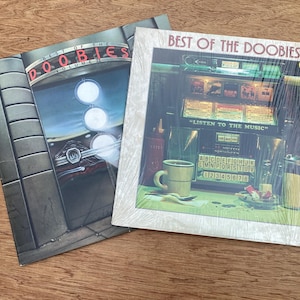 May include: Two vinyl record albums titled Best of the Doobies. One cover features a neon sign and car front, while the other shows a jukebox with the text Listen to the Music, a coffee mug, and a table setting. Both are square cardboard sleeves.