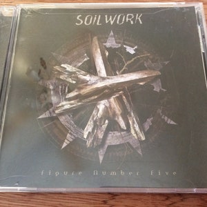 Może przedstawiać: Etui na płytę CD z ciemnozielonym tłem, na kt&oacute;rym w środku znajduje się drewniany obiekt w kształcie krzyża. Tekst "SOILWORK" jest nadrukowany na g&oacute;rze etui na biało. Tekst "figure number five" jest nadrukowany na dole etui na biało.