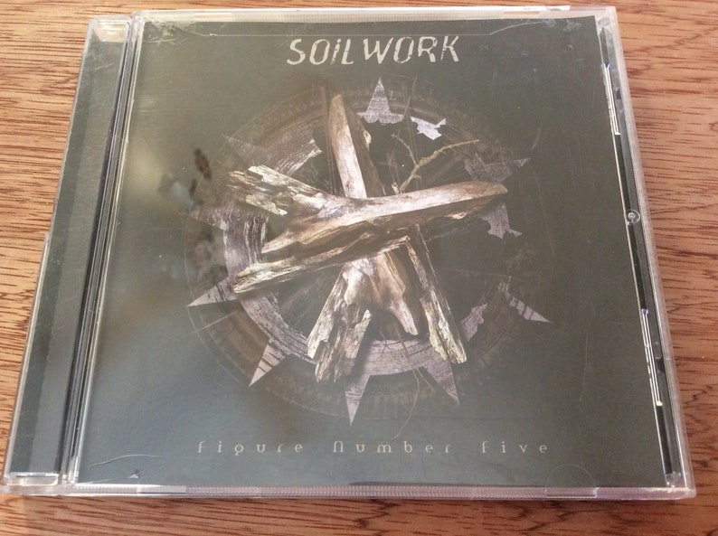 Może przedstawiać: Etui na płytę CD z ciemnozielonym tłem, na kt&oacute;rym w środku znajduje się drewniany obiekt w kształcie krzyża. Tekst "SOILWORK" jest nadrukowany na g&oacute;rze etui na biało. Tekst "figure number five" jest nadrukowany na dole etui na biało.