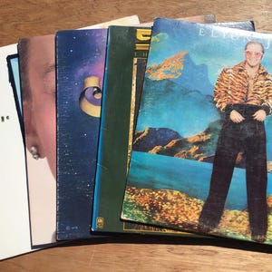 May include: A stack of vintage vinyl records. Album covers feature various designs, including a portrait, a starry sky, and an image of Elton John in a tiger-striped jacket. The records are arranged on a wooden surface, showcasing a collection of music.
