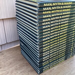 Puede incluir: Una pila de libros de tapa dura con el t&iacute;tulo "MAN, MYTH & MAGIC" en letras doradas sobre una cubierta verde oscuro. Los libros est&aacute;n apilados sobre una superficie de madera junto a una pared clara y texturizada.