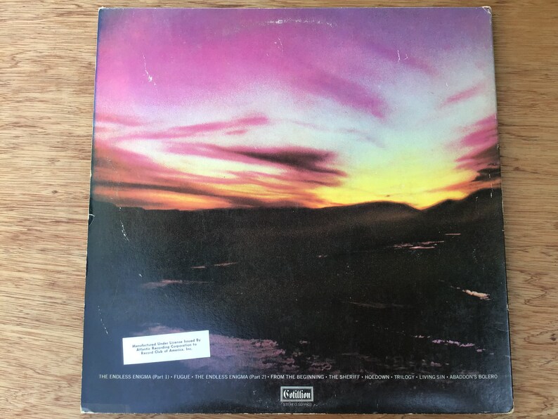 May include: The back cover of a vinyl record featuring a sunset over a mountain range. The record is titled "The Endless Enigma" and includes the tracks "From The Beginning", "The Sheriff", "Hoedown", "Trilogy", "Living Sin", and "Abaddon's Bolero".