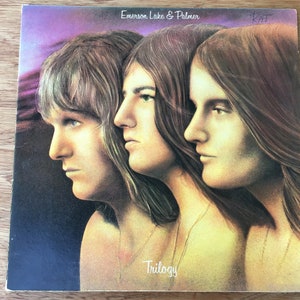 May include: A vintage vinyl record album cover featuring a photo of three men with long hair. The album is titled "Trilogy" by Emerson, Lake & Palmer.