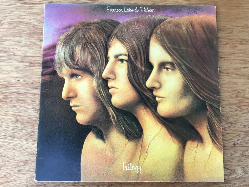 May include: A vintage vinyl record album cover featuring a photo of three men with long hair. The album is titled "Trilogy" by Emerson, Lake & Palmer.