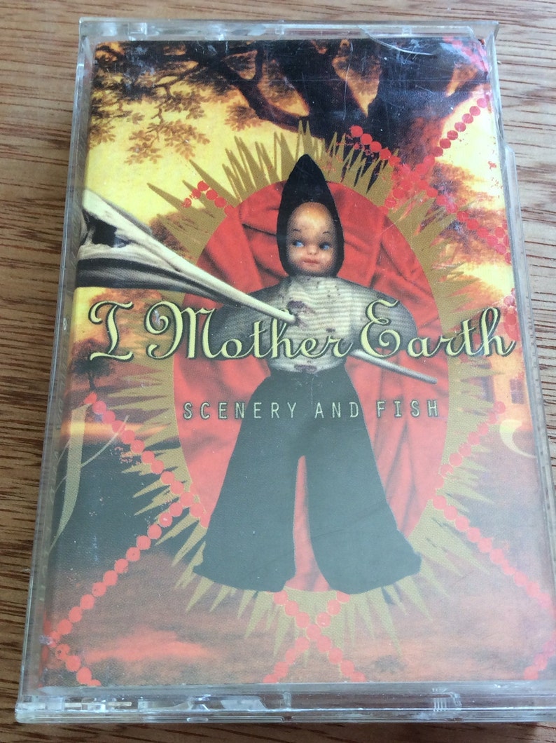 May include: A cassette tape with a red, gold and white design. The image features a doll with a striped shirt and black trousers. The text on the tape reads "I Mother Earth" and "Scenery and Fish".