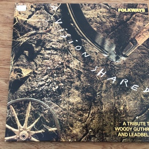 May include: Album cover for "A Vision Shared," a tribute to Woody Guthrie and Leadbelly. The cover features a textured, earthy background with a wagon wheel and tools. The title is in white, with the Folkways logo.