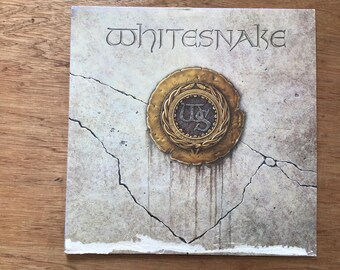 May include: Whitesnake album cover. The band's name is carved into the stone-like surface. A gold emblem with the initials "WS" is central. The background has a cracked, aged appearance. The design evokes a classic rock album.
