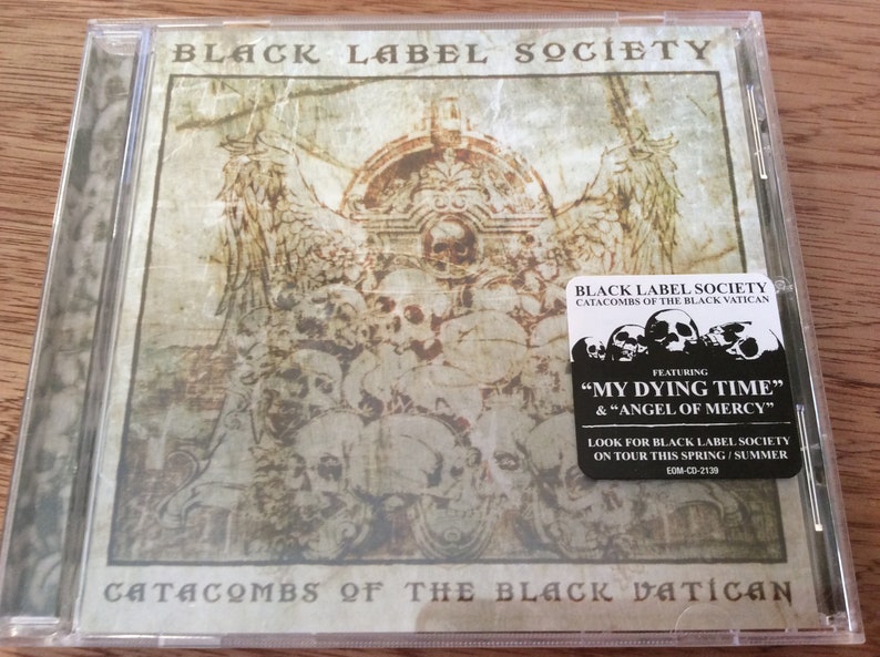 Może przedstawiać: Pudełko na płytę CD z czarno-białym obrazkiem czaszki i skrzyżowanych kości. Tekst na pudełku brzmi "Black Label Society Catacombs of the Black Vatican".