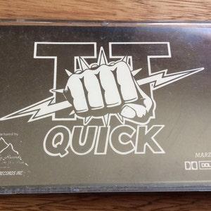 May include: A black cassette tape with a white graphic of a fist with a lightning bolt and the text "TT QUICK". The text "MARZT 2002" and "DOLBY SYSTEM" are printed on the bottom right corner of the tape.