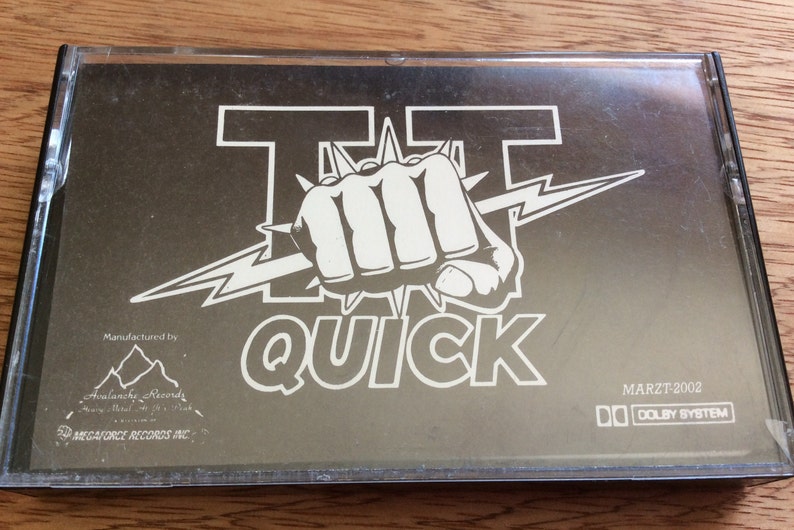 May include: A black cassette tape with a white graphic of a fist with a lightning bolt and the text "TT QUICK". The text "MARZT 2002" and "DOLBY SYSTEM" are printed on the bottom right corner of the tape.