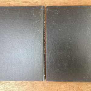 Puede incluir: Dos libros de tapa dura con cubiertas de color gris oscuro se muestran uno al lado del otro sobre una superficie de madera. Los libros tienen un aspecto ligeramente texturizado. Los lomos son visibles y las p&aacute;ginas est&aacute;n encuadernadas. Los libros son probablemente para leer o coleccionar.