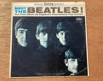 Puede incluir: Una funda de disco de vinilo vintage para Meet The Beatles! The First Album by England's Phenomenal Pop Combo. La portada presenta un retrato en blanco y negro de alto contraste de los cuatro miembros del grupo con texto azul.