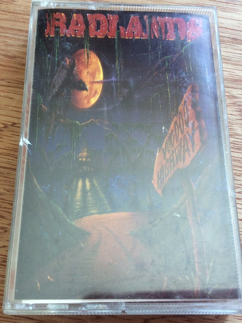 May include: A cassette tape featuring a dark, atmospheric image of a swamp with a full moon and a sign reading "Voodoo Highway". The band name "RADIANTS" is printed in red at the top of the cassette tape.
