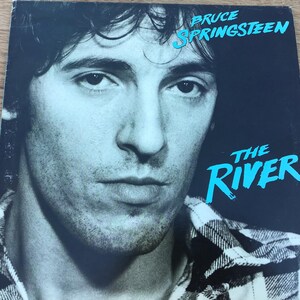 May include: A black and white photo of Bruce Springsteen on a vinyl record cover. The album cover is for "The River" album. The photo shows Springsteen looking directly at the camera with a serious expression. He is wearing a plaid shirt.