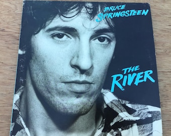 May include: A black and white photo of Bruce Springsteen on a vinyl record cover. The album cover is for "The River" album. The photo shows Springsteen looking directly at the camera with a serious expression. He is wearing a plaid shirt.