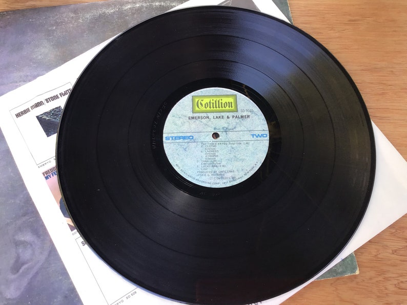 May include: Black vinyl record with a blue and white label. The label says "Cotillion" and "Emerson, Lake & Palmer". The record is labelled "Two" and lists the songs "The Three Fates", "Knife Edge", "Lucky Man", "Still... You Turn Me On", "From The Beginning", "Jeremy Bender", "Take A Pebble", "The Endless Enigma", "Close To Home", "The Battle Of Epping Forest", "Hoedown", "Lucky Man (Reprise)", "The Endless Enigma (Reprise)", "From The Beginning (Reprise)".