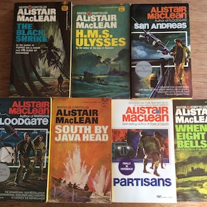 May include: A collection of vintage paperback books by Alistair MacLean, featuring titles like "The Black Shrike," "H.M.S. Ulysses," and "San Andreas." The covers display illustrations of action scenes and maritime themes, with text in bold, eye-catching fonts.