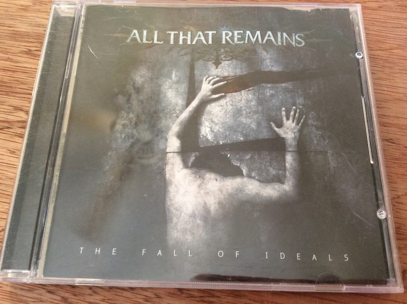 Może przedstawiać: Pudełko na płytę CD z czarno-białym obrazkiem osoby trzymającej duży kamienny blok. Tekst "ALL THAT REMAINS" znajduje się na g&oacute;rze pudełka, a "THE FALL OF IDEALS" na dole.