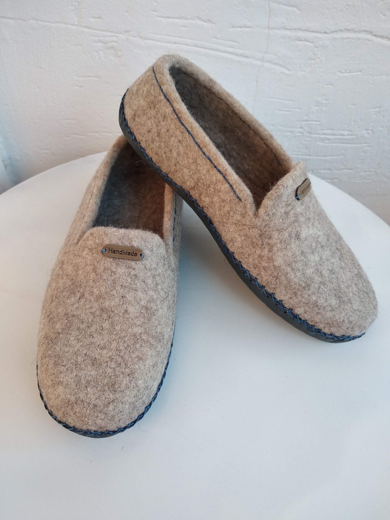 May include: A pair of gray felt slippers with a blue trim and a black sole. The slippers have a label that says "Handmade".