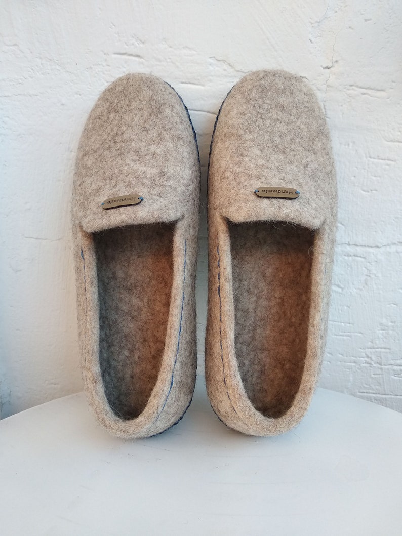 May include: A pair of gray wool slippers with blue stitching and a brown leather tag that says "Handmade".