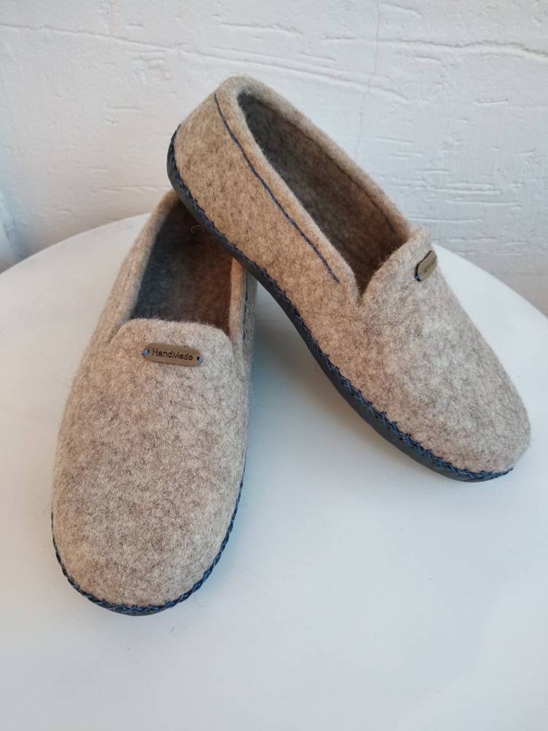 May include: A pair of gray felt slippers with a black trim and a blue stitching detail. The slippers have a label that says "Handmade".