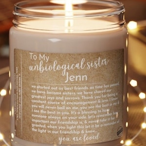 May include: A brown glass candle jar with a label that reads "To My Unbiological Sister Jenn". The label has a heartfelt message about the importance of friendship and sisterhood. The candle is sitting on a wooden coaster with a string of white lights around it.