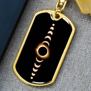 May include: A gold-colored dog tag keychain with a black background. The tag features a graphic of a solar eclipse, with phases of the moon. The keychain is attached to a gold-colored key ring and a key.