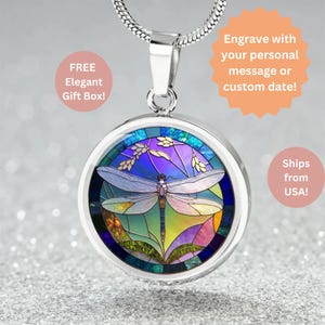 May include: Silver-tone pendant necklace featuring a round pendant with a stained-glass style dragonfly design. The dragonfly is light blue with colorful wings. The pendant hangs from a silver chain. The text "Engrave with your personal message or custom date!" is visible.