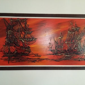May include: A framed painting depicting a seascape with several ships silhouetted against a fiery orange sunset. The ships are painted in black with gold accents, and the water is a dark blue.