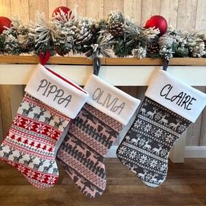 May include: Three personalised Christmas stockings with names embroidered on the white cuffs. The stockings feature festive patterns with reindeer and snowflakes in red, pink, and grey. A garland with red baubles and pinecones decorates the background.