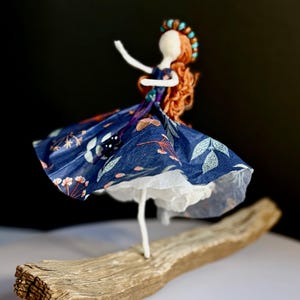 May include: A handmade doll with a red yarn hairstyle and a blue floral dress. The doll is standing on a piece of driftwood. The doll's head is white, and it has a turquoise beaded headband. The dress has a white underskirt.