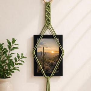 May include: A sage green macrame wall hanging featuring a wooden ring at the top, intricate knotted cord patterns, and a central black rectangular picture frame displaying a desert landscape with a saguaro cactus at sunset, finished with a tassel.