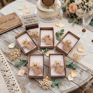 May include: Four rectangular white soap bars in brown cardboard boxes, each decorated with twine, dried baby's breath flowers, and a kraft paper tag reading Alexander & Charlotte 18.07.2027. A wooden guest book and a sign reading Welcome to Our Celebration.
