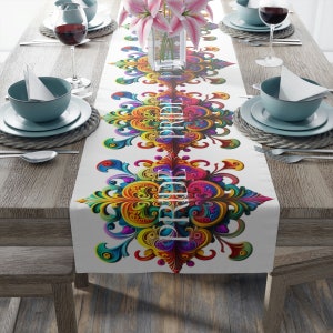 Puede incluir: Un camino de mesa blanco con un diseño simétrico y vibrante en tonos de amarillo, naranja, rosa, verde y azul. El diseño presenta patrones ornamentados y arremolinados. La palabra "BRIGHT" está impresa verticalmente en blanco. El camino de mesa está sobre una mesa de madera.