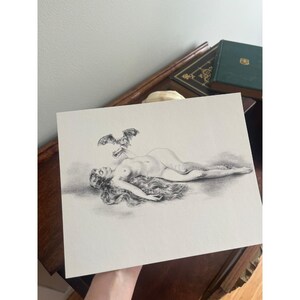 May include: A pencil drawing on white paper shows a nude figure reclining with long flowing hair. Above the figure, a bat is drawn. The artwork is held by a hand, with a glimpse of a wooden surface and books in the background.