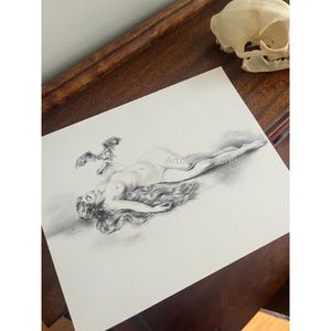 May include: A pencil drawing of a reclining figure with long flowing hair and a small bat-like creature. The artwork is on white paper, with the artist's name visible in the corner. A cat skull rests on a wooden surface.