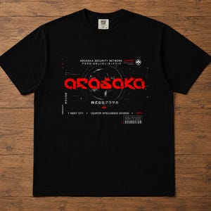 May include: A black short-sleeve t-shirt featuring a graphic print with the red text ARASAKA in a stylized font. The design includes white text reading ARASAKA SECURITY NETWORK, NIGHT CITY, COUNTER INTELLIGENCE DIVISION, and 2077, along with Japanese characters.