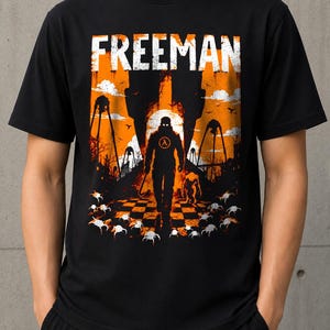 Puede incluir: Una camiseta negra de manga corta con un estampado gráfico que incluye el texto FREEMAN en la parte superior. El diseño muestra la silueta de una figura con una palanca sobre un fondo naranja con estructuras altas, criaturas y un suelo de damero.