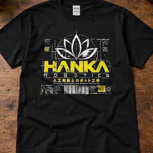 Puede incluir: Una camiseta negra de manga corta con un estampado gráfico de una flor de loto blanca sobre el texto amarillo HANKA ROBOTICS. Debajo, aparecen caracteres japoneses y el texto ARTIFICIAL INTELLIGENCE AND ROBOTICS ENGINEERING.