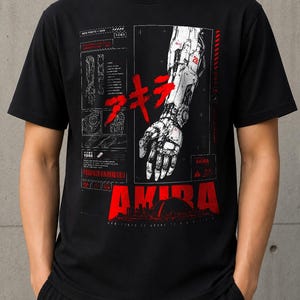Op de afbeelding: Een zwart t-shirt met korte mouwen en een grafische print van een mechanische robotarm in wit en grijs. Het ontwerp bevat rode Japanse tekst, het woord AKIRA in dikke rode letters en technische schema's met kleine witte tekstdetails.