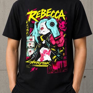 Può includere: Una t-shirt nera a maniche corte con una stampa grafica del personaggio Rebecca di Cyberpunk Edgerunners. Il design include il testo REBECCA, CYBERPUNK EDGERUNNERS e caratteri giapponesi, con vivaci accenti color rosa neon, giallo e blu.