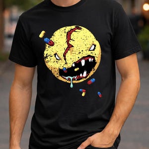 Puede incluir: Camiseta negra con un diseño de cara sonriente amarilla desgastada, con una cicatriz roja y dientes afilados. Pastillas de colores dispersas alrededor de la cara. El diseño tiene una estética grunge.