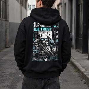 Op de afbeelding: Een zwarte hoodie met een grafische print op de achterkant. Het ontwerp toont een soldaat met een gasmasker, stadssilhouetten en de tekst IN THE COMBINE WE TRUST in lichtblauw, aangevuld met technische gegevens en diverse symbolen.