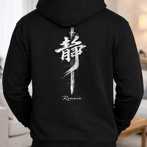 May include: Black hooded sweatshirt with a white graphic design on the back. The design features a stylized sword and Japanese characters, with the word "Remain" below. The sweatshirt has a drawstring hood and a kangaroo pocket.
